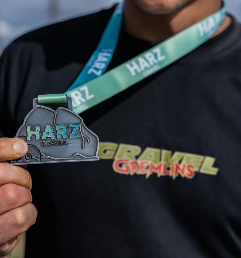 Person hält eine Medaille mit der Aufschrift "HARZ" und trägt ein schwarzes T-Shirt mit buntem Schriftzug.