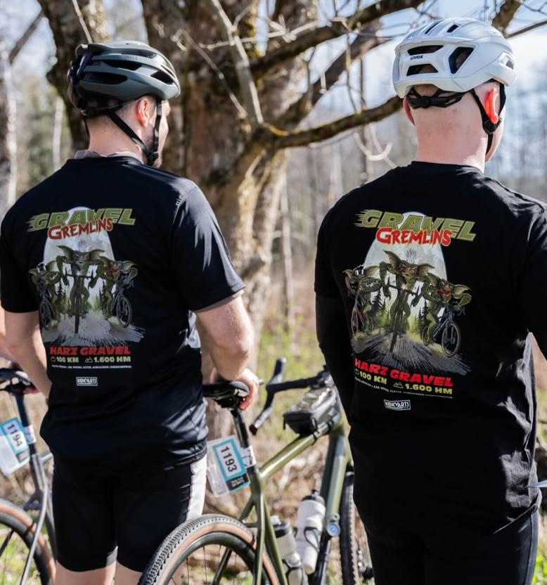 Drei Personen mit Fahrradhelmen stehen neben Fahrrädern im Freien und tragen schwarze T-Shirts mit buntem Aufdruck auf dem Rücken.
